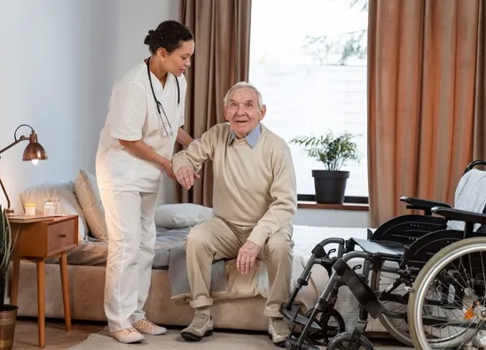 Professional caregiver helping senior resident stand beside wheelchair in assisted living facility
