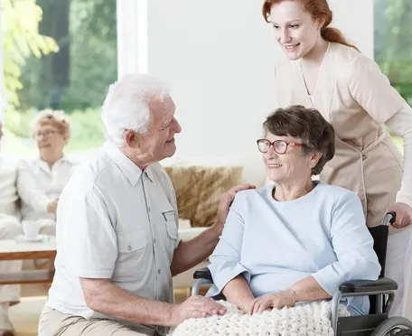 Caregiver assisting senior woman in wheelchair while elderly couple talks in assisted living community
